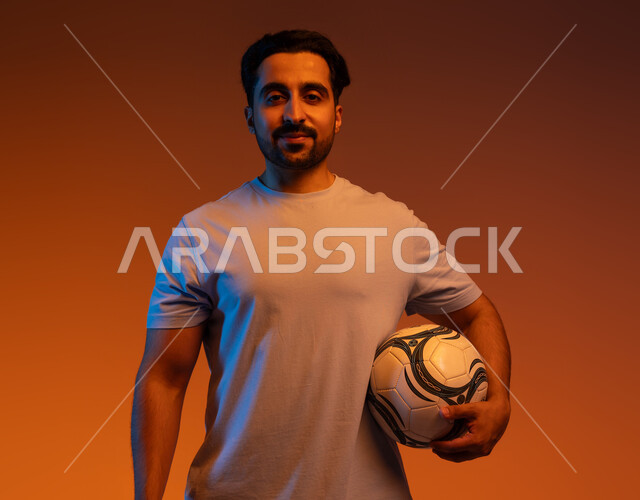 الاستعداد لبدء المباراة، الوقوف باستقامة والنظر إلى الكاميرا، ممارسة الهواية المفضلة، بورتريه مقرب للاعب عربي سعودي خليجي يرتدي الزي الكاجوال يحمل كرته بيده، احتراف الرياضات الجماعية، احتراف اللعب بالكره القدم، التسلية و الترفيه بالرياضات الحركية، خلفية ملونة