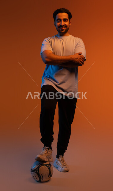 احتراف اللعب بكره القدم، النظر إلى الكاميرا بايماءات  تدل على الثقة، الاستعداد لبدء المباراة، ممارسة الهواية المفضلة، بورتريه للاعب عربي سعودي خليجي يرتدي الزي الكاجوال، احتراف الرياضات الجماعية، التسلية و الترفيه بالرياضات الحركية، صورة الجسم بالكامل، خلفية ملونة