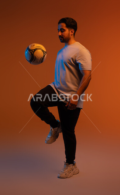 احتراف الرياضات الجماعية، النظر إلى الكاميرا بايماءات  تدل على الثقة، الاستعداد لبدء المباراة، ممارسة الهواية المفضلة، بورتريه للاعب عربي سعودي خليجي يرتدي الزي الكاجوال، احتراف اللعب بكره القدم، التسلية و الترفيه بالرياضات الحركية، صورة الجسم بالكامل، خلفية ملونة