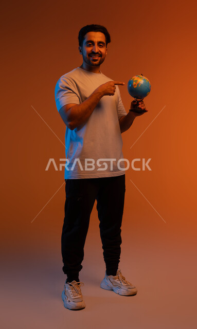 الوقوف باستقامة و النظر الى الكاميرا، تعابير السرور بالانتماء للدول العربيه, البحث واستكشاف القارات في الجغرافيا , الاشاره الى مجسم الكره الارضيه,  بورتريه لرجل عربي سعودي خليجي يرتدي الزي الكاجوال يشير الى خريطة العالم, صورة لكامل الجسم، خلفيه ملونه