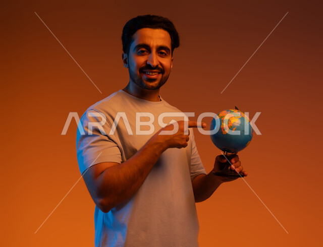 Expressions of pleasure in belonging to Arab countries, research and exploration of continents in geography, reference to a globe, portrait of a Saudi Gulf Arab man wearing casual clothing pointing to the world map, colored background