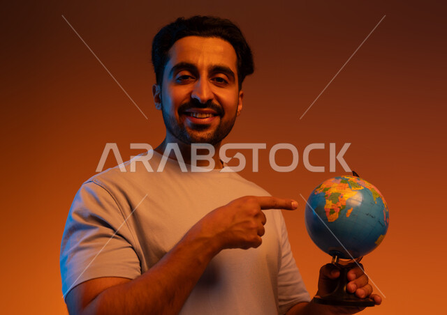 البحث واستكشاف القارات في الجغرافيا , الاشاره الى مجسم الكره الارضيه,  بورتريه مقرب لرجل عربي سعودي خليجي يرتدي الزي الكاجوال يشير الى خريطة العالم, تعابير السرور بالانتماء للدول العربية, خلفيه ملونه