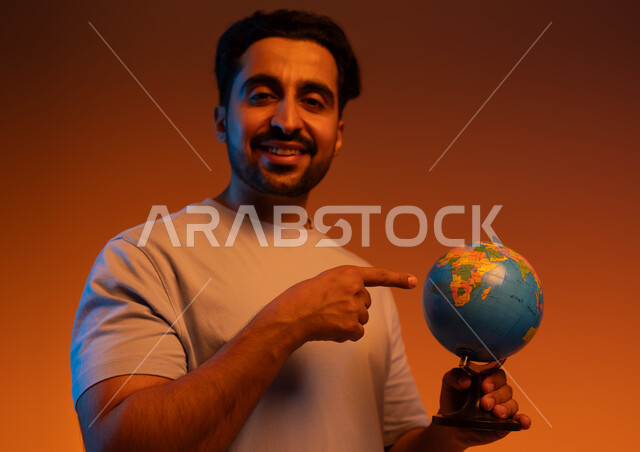 الاشاره الى مجسم الكره الارضيه,  بورتريه مقرب لرجل عربي سعودي خليجي يرتدي الزي الكاجوال يشير الى خريطة العالم, تعابير السرور بالانتماء للدول العربيه, البحث واستكشاف القارات في الجغرافيا , خلفيه ملونه