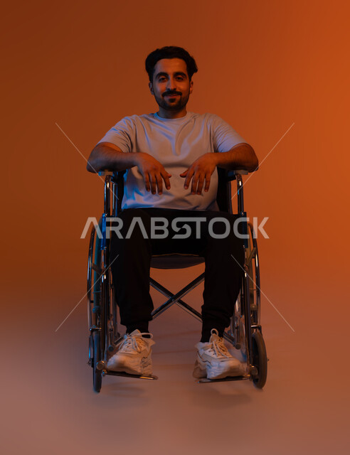 Weakness in motor ability, looking at the camera with gestures of optimism and hope, a portrait of a young Arab, Saudi, Gulf, seated in casual uniform, sitting in a wheelchair, providing supplies for the disabled, colorful background