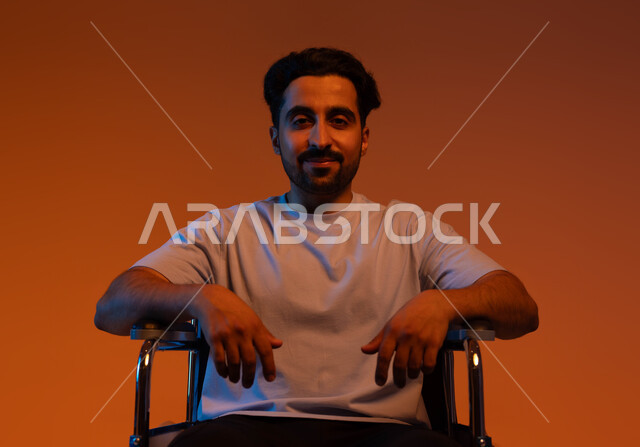 Sitting in a wheelchair, portrait of a crippled young Arab Saudi Gulf man wearing casual clothing looking at the camera with gestures of optimism and hope, providing special supplies for the disabled, colorful background