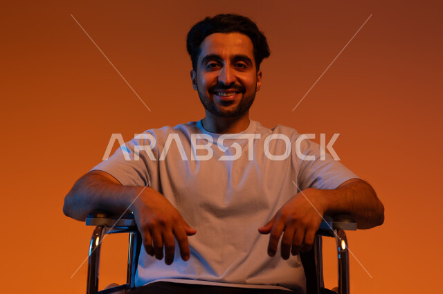 مفهوم الإعاقة الحركية،  النظر إلى الكاميرا بإيماءات الأمل  والتفاؤل , بورتريه مقرب لشاب عربي سعودي خليجي مقعد يرتدي الزي الكاجوال يجلس على كرسي متحرك, توفير المستلزمات الخاصه بالمعاقين , خلفيه ملونه