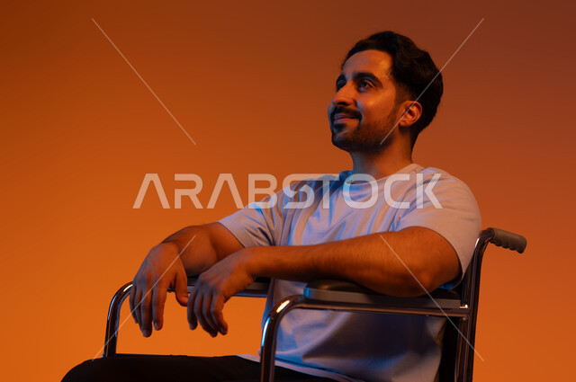توفير المستلزمات الخاصه بالمعاقين, ايماءات التأمل والتفاؤل, بورتريه مقرب لشاب عربي سعودي خليجي مقعد يرتدي الزي الكاجوال يجلس على كرسي متحرك, النظر الى شيء ما, خلفيه ملونه