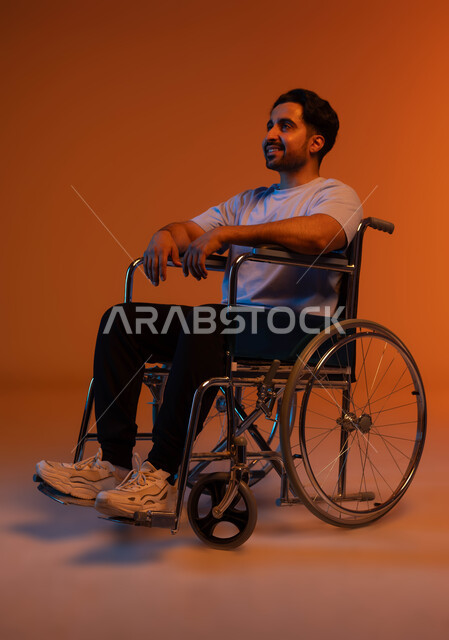 شعور المعاقين بالإنتماء والتقدير والإحترام في السعوديه، تأمين مستلزمات تساعد على الحركة والتنقل، بورتريه لشاب عربي سعودي خليجي مبتسم يرتدي الزي الكاجوال يجلس على كرسي متحرك وبنظر الى شيء ما بإيماءات السرور، خلفيه ملونة .