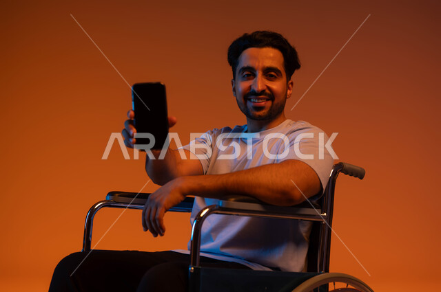 Using advanced modern technology, a portrait of a smiling, seated Saudi Arabian Gulf man wearing a casual outfit, displaying a blank black screen on his mobile phone and looking at the camera with happy gestures, a colored background.