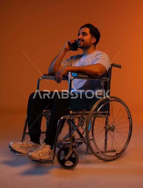 Laughing and hanging out with friends, making a voice call on the mobile phone, paying attention to the psychology of people of determination and giving them positive energy, communicating and chatting with relatives, a portrait of a crippled Saudi Gulf Arab man wearing a casual outfit sitting in a wheelchair and holding a mobile phone in his hand, a colorful background.