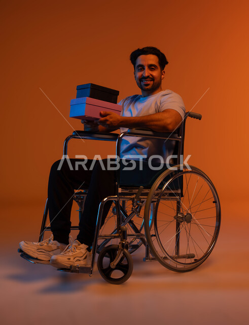 شعور المعاقين بالحب و التقدير، اهتمام ودعم المجتمع السعودي لذوي الهمم, مناسبات و أعياد سعيدة، بورتريه لشاب عربي خليجي سعودي مقعد يرتدي الزي الكاجوال يجلس على كرسي متحرك و يمسك بيديه علب الهدايا الملونة، النظر الى الكاميرا بإيماءات السرور, خلفية ملونة .