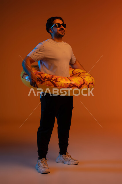 قضاء عطلة صيفيه ممتعه ،رحلة سياحية ترفيهيه، النظر إلى شيء ما بايماءات السرور ،بورتريه لشاب عربي خليجي سعودي يرتدي الزي الكاجوال والنظارات الشمسية و عوامة السباحة، مفهوم التسلية في أوقات الفراغ، صورة الجسم بالطول الكامل ،خلفية ملونة .