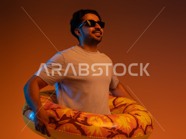 . A recreational tourist trip, looking at something with gestures of pleasure and happiness, spending an enjoyable summer vacation, a close-up portrait of a young Saudi Gulf Arab man wearing casual clothes, sunglasses and a swimming buoy, the concept of leisure time entertainment, colorful background.