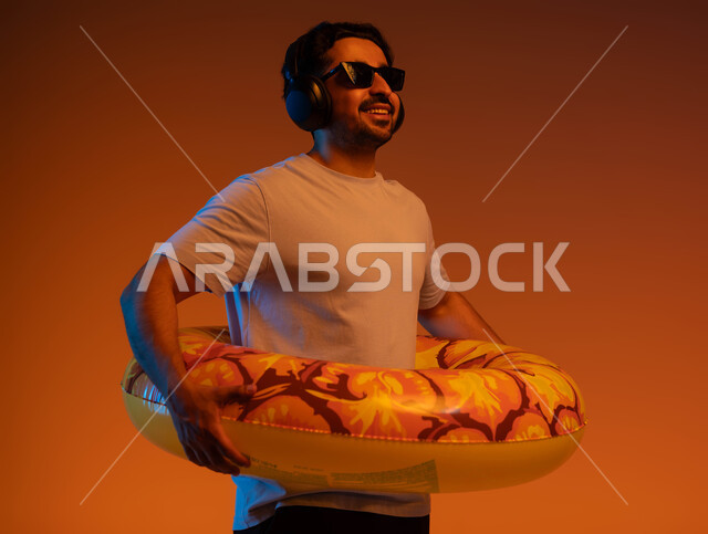 استخدام التقنيات الحديثة المتطورة، الاستعداد لقضاء عطلة صيفيه ،رحلة سياحية ممتعه، بورتريه مقرب لشاب عربي خليجي سعودي يرتدي الزي الكاجوال و سماعات الهيدفون و عوامة السباحة، مفهوم الترفيه و التسلية ،النظر إلى شيء ما بايماءات السرور، خلفية ملونة .