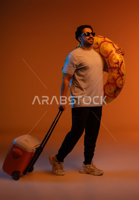 . Preparing for the summer vacation with gestures of pleasure, a fun tourist trip, a portrait of a Saudi Gulf Arab young man wearing a casual outfit and sunglasses, carrying a swimming buoy on his shoulder and holding an ice box in his hand, the concept of leisure and entertainment, full body portrait, colorful background