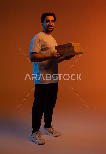 خدمات تسليم البضائع الأونلاين للزبائن، مفهوم التسوق الإلكتروني الآمن, بورتريه مقرب لمندوب توصيل عربي خليجي سعودي مبتسم يحمل بيده علب كرتونية و يقوم بتوصيل الطلبات السريعه ،النظر إلى الكاميرا بإيماءات السرور، صورة الجسم بالكامل، خلفية ملونة .