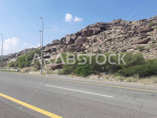 الطرقات الاسفلتية المعبدة في أماكن القمم والمرتفعات الجبلية في وقت النهار، طريق بين مدينة الحجرة في منطقة الباحة ومدينة أضم في مكة المكرمة، منظر الغيوم الكثيفة في السماء الزرقاء، النباتات الخضراء البرية على حافتي الشارع، أعمدة الإنارة الطرقية