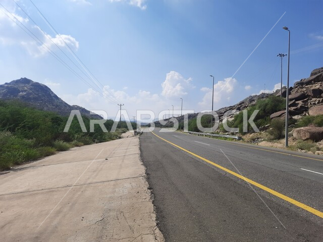 الطرقات الاسفلتية المعبدة في أماكن القمم والمرتفعات الجبلية في وقت النهار، طريق بين مدينة الحجرة في منطقة الباحة ومدينة أضم في مكة المكرمة، منظر الغيوم الكثيفة في السماء الزرقاء، النباتات الخضراء البرية على حافتي الشارع، أعمدة الإنارة الطرقية