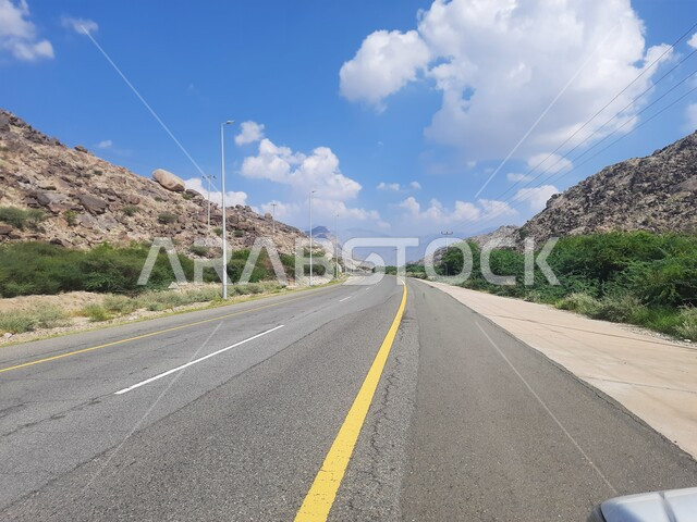 الطرقات الاسفلتية المعبدة في أماكن القمم والمرتفعات الجبلية في وقت النهار، طريق بين مدينة الحجرة في منطقة الباحة ومدينة أضم في مكة المكرمة، منظر الغيوم الكثيفة في السماء الزرقاء، النباتات الخضراء البرية على حافتي الشارع، أعمدة الإنارة الطرقية