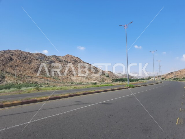 الطرقات الاسفلتية المعبدة في أماكن القمم والمرتفعات الجبلية في وقت النهار، طريق بين مدينة الحجرة في منطقة الباحة ومدينة أضم في مكة المكرمة، منظر الغيوم الكثيفة في السماء الزرقاء، النباتات الخضراء البرية على حافتي الشارع، أعمدة الإنارة الطرقية