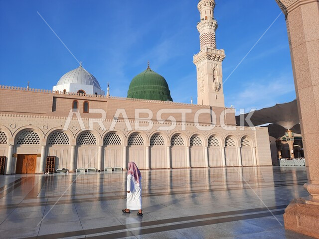 التقرب الى الله تعالى بزيارة المعالم الدينية الإسلامية، صورة من الخلف لرجل عربي خليجي سعودي يمشي في الحرم النبوي الشريف، القبة الخضراء ومئذنة المسجد النبوي في المدينة المنورة بالمملكة العربية السعودية