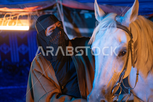 ارتداء أزياء يوم التأسيس السعودي 1727، ذكرى يوم التاسيس السعودي 22 فبراير، ملاطفة الخيول والاهتمام بهم، صورة مقربة لإمرأة عربية خليجية سعودية ترتدي العباءة والبرقع تقف بجانب فرسها الأبيض في وقت الليل، كشتة بر بدوية، مشاعر الحب والعشق للخيل السعودي