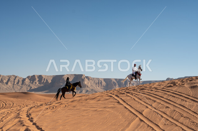 قوة وأصالة الحصان العربي، الاحتفال بالمناسبات والأعياد الوطنية، فعاليات سباق الخيول العربية في الصحراء، فارسان عربيان خليجيان سعوديان يرتديان الزي التقليدي يمتطيان الجياد ويتسابقان فوق الرمال الذهبية في وضح النهار