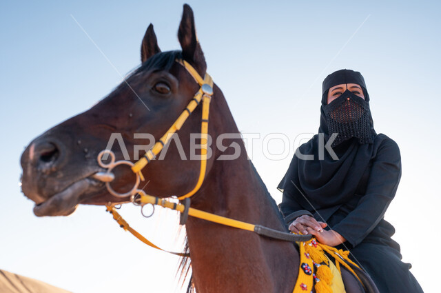 Establishment of the first Saudi state February 22, petting and caring for horses, horse riding activities and events, a close-up of a Gulf Arab woman wearing a burqa and abaya riding her brown horse in the desert in broad daylight, practicing the hobby of horse riding, a love of equestrianism and riding horses.