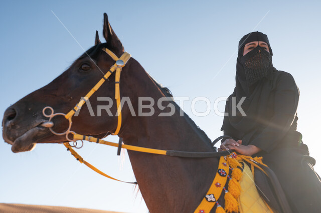 حب الفروسيه وركوب الجياد، تأسيس الدولة السعودية الأولى 22 فبراير، ملاطفة الخيول والاعتناء بهم، أنشطة وفعاليات ركوب الأحصنة، صورة مقربة لإمرأة عربيه خليجية ترتدي البرقع والعباءة تمتطي فرسها البني في الصحراء في وضح النهار ، ممارسة هواية ركوب الخيل