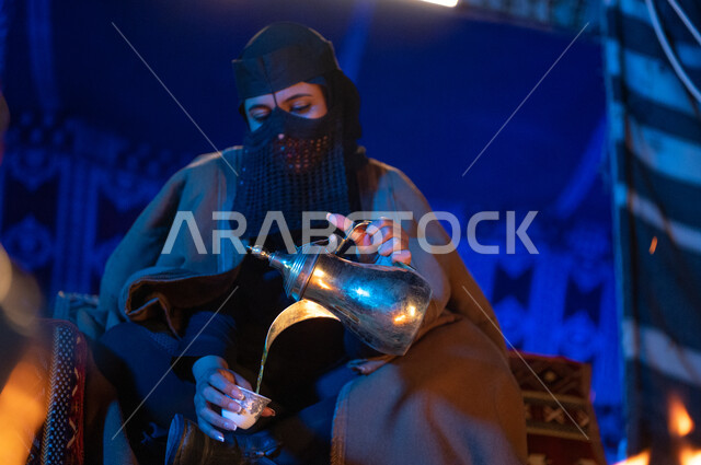 تقاليد وعادات سعودية اصيلة في تقديم القهوة العربية، مفهوم الكرم وحسن الضيافه والاستقبال، صورة مقربة لإمرأة عربية خليجية سعودية ترتدي العباءة الفرو والبرقع تقوم بصب القهوة باستخدام الدلة النحاسية في فناجين داخل خيمة شعبية تراثية، منتج محلي ومحاصيل زراعية وطنية