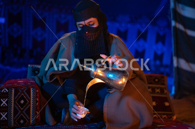 عادات وتقاليد سعودية اصيلة في تقديم القهوة العربية، مفهوم الكرم وحسن الضيافه والاستقبال، صورة مقربة لإمرأة عربية خليجية سعودية ترتدي العباءة الفرو والبرقع تقوم بصب القهوة باستخدام الدلة النحاسية في فناجين داخل خيمة شعبية تراثية، منتج محلي ومحاصيل زراعية وطنية