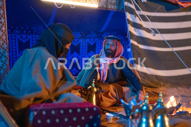 قضاء أوقات عائلية ممتعة في رحلة في الصحراء السعودية، مفهوم الكرم وحسن الضيافه والاستقبال، صورة مقربة لزوجين عربيين خليجيين سعوديين يرتديان الفروة يجلسان داخل خيمة شعبية تراثية ويستمتعان بشرب القهوة العربية، كشتة بر وتخييم في الأجواء الشتوية 