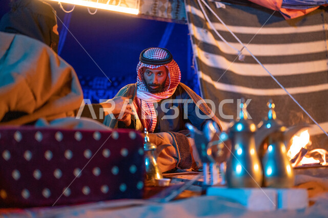 قضاء أوقات عائلية ممتعة في رحلة في الصحراء السعودية، مفهوم الكرم وحسن الضيافه والاستقبال، صورة مقربة لزوجين عربيين خليجيين سعوديين يرتديان الفروة يجلسان داخل خيمة شعبية تراثية ويستمتعان بشرب القهوة العربية، كشتة بر وتخييم في الأجواء الشتوية 