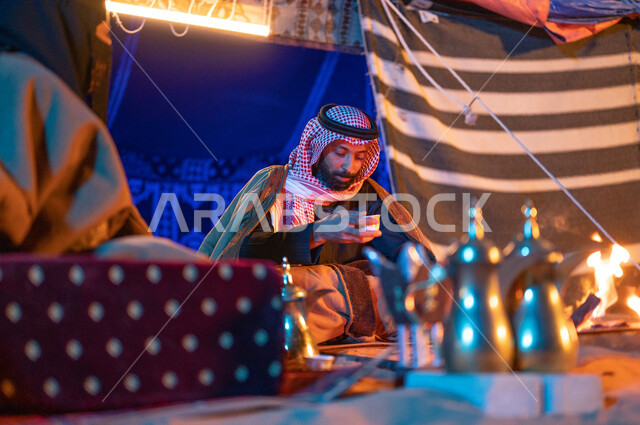 قضاء أوقات عائلية ممتعة في رحلة في الصحراء السعودية، مفهوم الكرم وحسن الضيافه والاستقبال، صورة مقربة لزوجين عربيين خليجيين سعوديين يرتديان الفروة يجلسان داخل خيمة شعبية تراثية ويستمتعان بشرب القهوة العربية، كشتة بر وتخييم في الأجواء الشتوية 