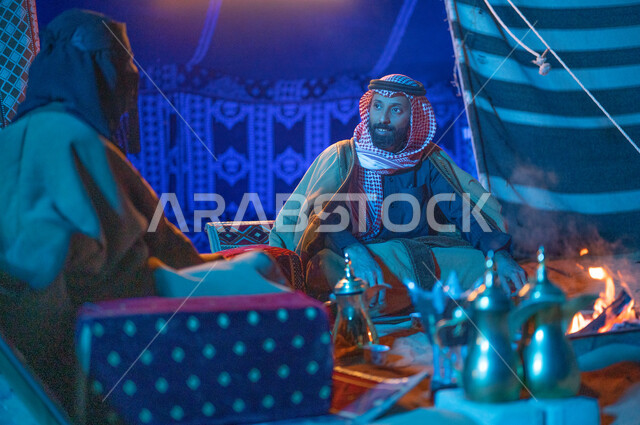 مفهوم الكرم وحسن الضيافه، كشتة بر وتخييم في الأجواء الشتوية، قضاء أوقات عائلية ممتعة في رحلة في الصحراء السعودية، صورة مقربة لزوجين عربيين خليجيين سعوديين يرتديان الفروة يجلسان داخل خيمة شعبية تراثية ويستمتعان بشرب القهوة العربية