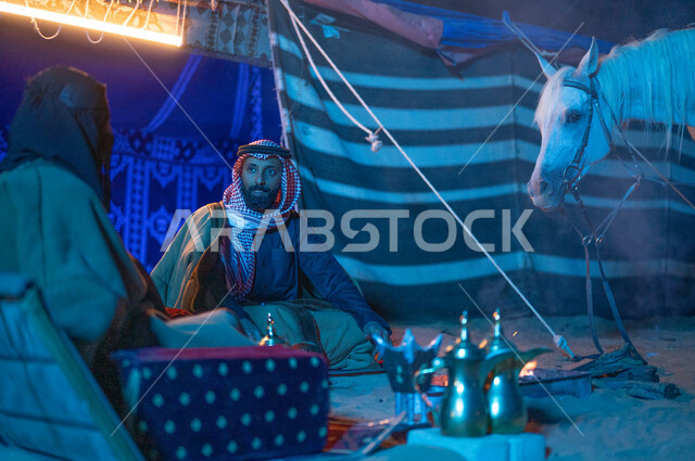 ارتداء زي يوم التأسيس السعودي الشعبي، كشتة بر وتخييم في الأجواء الشتوية بصحبة الخيل الأصيل، قضاء أوقات عائلية ممتعة في الصحراء السعودية، صورة مقربة لزوجين عربيين خليجيين سعوديين يرتديان الفروة يجلسان داخل خيمة شعبية تراثية ويستمتعان بشرب القهوة العربية