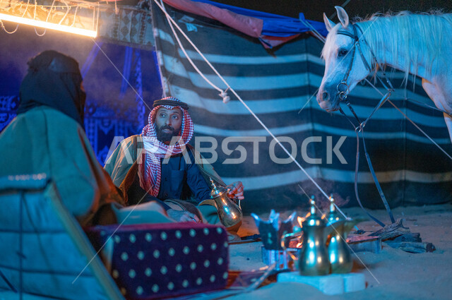 ارتداء زي يوم التأسيس السعودي الشعبي، كشتة بر وتخييم في الأجواء الشتوية بصحبة الخيل الأصيل، قضاء أوقات عائلية ممتعة في الصحراء السعودية، صورة مقربة لزوجين عربيين خليجيين سعوديين يرتديان الفروة يجلسان داخل خيمة شعبية تراثية ويستمتعان بشرب القهوة العربية