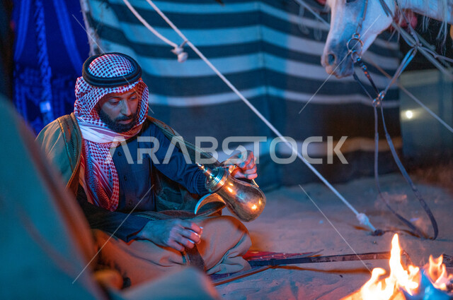 The concept of good hospitality and reception, the Saudis’ passion for raising horses, spending enjoyable times in the Saudi desert, camping in the winter, a Saudi Gulf Arab man sitting inside a popular traditional tent pouring Arabic coffee using a copper bucket.