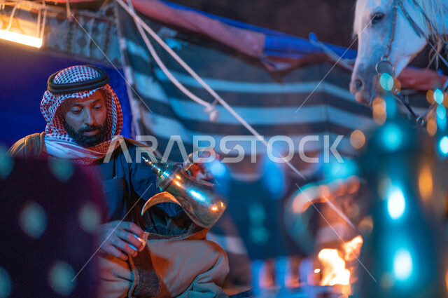 مفهوم حسن الضيافة والاستقبال، شغف السعوديين بتربية الجياد، قضاء أوقات ممتعة في الصحراء السعودية، كشتة بر وتخييم في الأجواء الشتوية، رجل عربي خليجي سعودي يجلس داخل خيمة شعبية تراثية يقوم يقوم بصب القهوة العربية باستخدام الدلة النحاسية