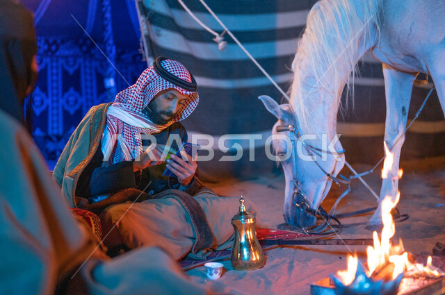 Spending fun time with the family in Kashta Barr and camping in the winter atmosphere, a Gulf man using a mobile phone, wearing the popular Saudi Founding Day costume, a Saudi Gulf Arab couple wearing furs sitting inside a popular tent in the desert, the Saudis’ passion for raising horses