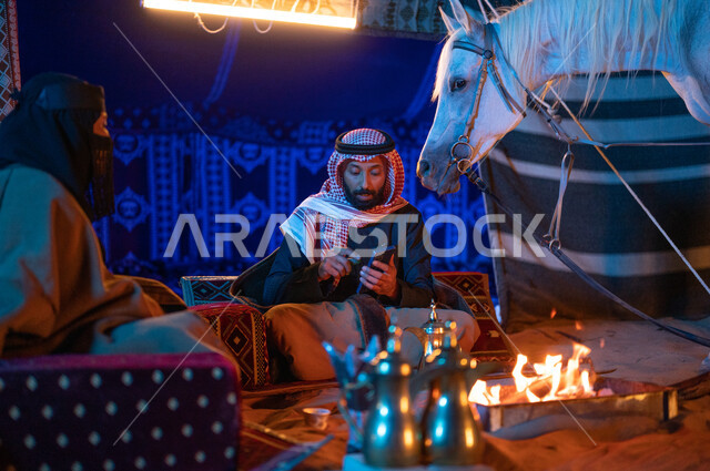 قضاء وقت ممتع مع العائلة في كشتة بر وتخييم في الأجواء الشتوية، رجل خليجي يقوم باستخدام الهاتف الجوال، ارتداء زي يوم التأسيس السعودي الشعبي، زوجان عربيان خليجيان سعوديان يرتديان الفروة يجلسان داخل خيمة شعبية في الصحراء، شغف السعوديين بتربية الخيول