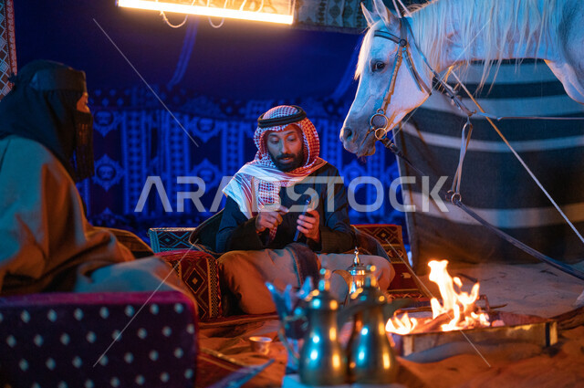  رجل خليجي يقوم باستخدام الهاتف الجوال، ارتداء زي يوم التأسيس السعودي الشعبي، قضاء وقت ممتع مع العائلة في كشتة بر وتخييم في الأجواء الشتوية، زوجان عربيان خليجيان سعوديان يرتديان الفروة يجلسان داخل خيمة شعبية في الصحراء، شغف السعوديين بتربية الخيول