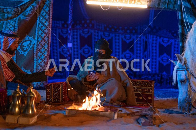 A Saudi woman wearing the popular Founding Day costume using a mobile phone, spending quality time with the family in Kashta Barr and camping in the winter atmosphere, a Saudi Gulf Arab couple wearing furs sitting inside a popular tent in the desert, the Saudis’ passion for raising horses
