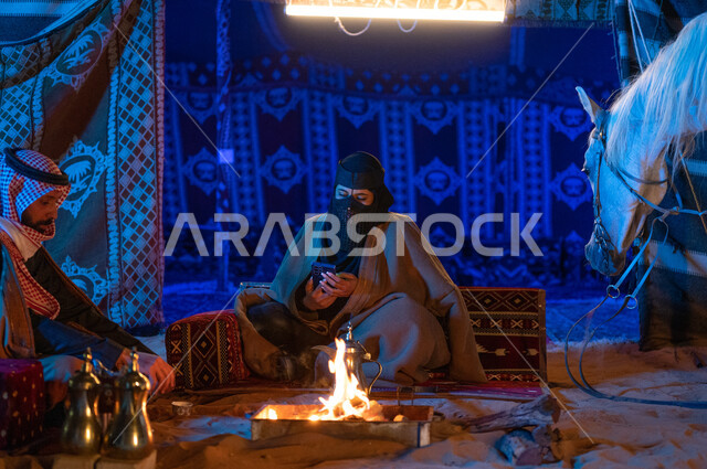 A Saudi woman wearing the popular Founding Day costume using a mobile phone, spending quality time with the family in Kashta Barr and camping in the winter atmosphere, a Saudi Gulf Arab couple wearing furs sitting inside a popular tent in the desert, the Saudis’ passion for raising horses