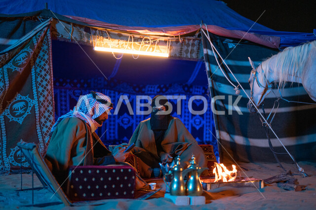  رجل خليجي يقوم باستخدام الهاتف الجوال، ارتداء زي يوم التأسيس السعودي الشعبي، قضاء وقت ممتع مع العائلة في كشتة بر وتخييم في الأجواء الشتوية، زوجان عربيان خليجيان سعوديان يرتديان الفروة يجلسان داخل خيمة شعبية في الصحراء، شغف السعوديين بتربية الخيول