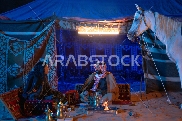 حب وشغف تربية الخيول الاصيلة، جلسة تخييم في الأجواء الشتوية في الصحراء السعودية، كشتة بر عائلية سعيدة، مفهوم الكرم وحسن الضيافه والاستقبال، صورة مقربة لزوجين عربيين خليجيين سعوديين يرتديان الفروة يجلسان داخل خيمة شعبية تراثية ويستمتعان بشرب القهوة العربية