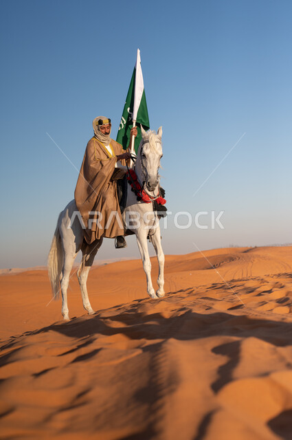 الاحتفال بذكرى تأسيس المملكة، مشاركة الجياد في المناسبات والأعياد الوطنية, رجل عربي خليجي سعودي يرتدي العقال العريض والبشت التقليدي يحمل علم التأسيس يقوم بامتطاء فرسه الابيض في وضح النهار في الصحراء, ارتداء أزياء أهل المنطقه الوسطى في يوم التأسيس 1727