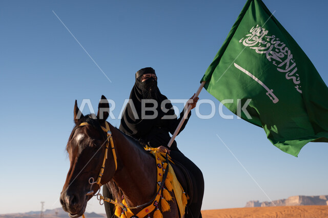 Raising the Saudi flag and flag in the desert, establishing the first Saudi state on February 22, horse riding activities and events, a Gulf Arab woman wearing a burqa and abaya riding her brown horse in broad daylight, practicing the hobby of horse riding, love of equestrianism and riding horses, celebrating Saudi Flag Day