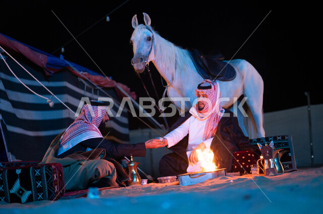 The concept of hospitality and good reception, popular sessions inside a heritage tent, authentic Saudi customs and traditions in serving Arabic coffee, a passion for raising purebred horses, two Saudi Gulf Arab friends wearing a shemagh and a bisht spending enjoyable times in a youthful kashta bar.