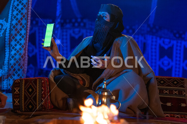 استخدام الوسائل التقنية الحديثة، إشعال موقد النار أثناء التخييم في الصحراء السعودية، كشتة بر في الأجواء الشتوية الهادئة، امرأة عربية خليجية سعودية ترتدي البرقع والفروة تقوم باستخدام الهاتف الجوال بشاشة كروما خضراء فارغة داخل خيمة شعبية تراثية